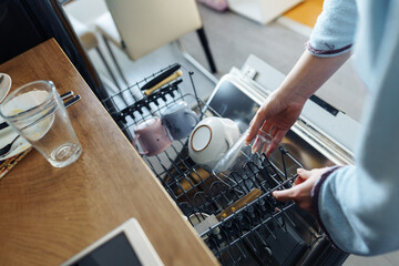 young woman putting dirty dishes into dishwasher at home