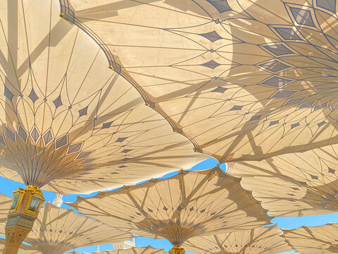 Low-angle view of the majestic retractable umbrellas providing shade in the courtyard of Al Masjid an Nabawi in Medina. The umbrellas feature intricate geometric patterns and golden columns.