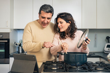 Authentic middle aged couple cooking together using online recipe on tablet in modern kitchen