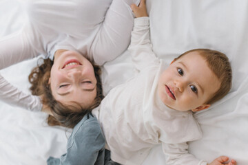 Happy mother and baby lying together on a soft white bed, enjoying a warm and cozy family moment filled with love, comfort, connection, and natural light.