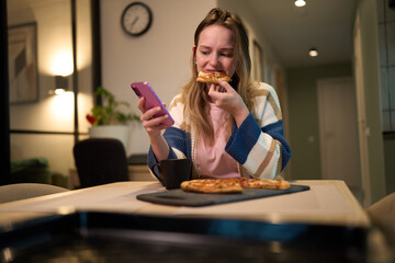 Female eating pizza while using smartphone at kitchen table