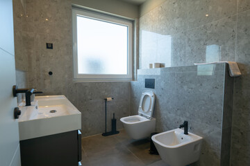 Modern, luxurious bathroom featuring a double vanity, sleek grey marble tiles, a walk-in shower, and a minimalist toilet. A mirror reflects a glimpse of the adjoining stylish bedroom