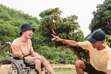 A wheelchair user relaxes at a campsite while a companion sets up a tent nearby. They enjoy accessible camping together, highlighting inclusion, teamwork, outdoor travel, positive lifestyle in nature.