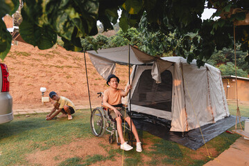 A wheelchair user relaxes at a campsite while a companion sets up a tent nearby. They enjoy accessible camping together, highlighting inclusion, teamwork, outdoor travel, positive lifestyle in nature.
