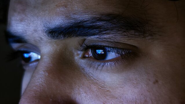 close up detail of focused male eyes staring at computer monitor screen with blue light reflection in dark night