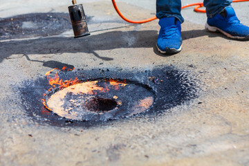worker uses a propane torch to heat roofing material around a circular patch, creating a charred, flame-active zone. This high-temperature process is part of roof surface sealing or repair