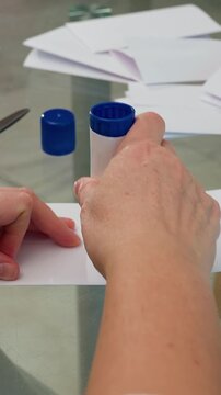 glue stick folded paper white woman assembling small booklets on glass table, scissors and stacked envelopes visible, careful finger creasing, teacher preparing flashcards, event planner prepping