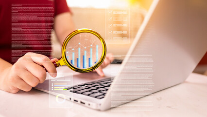 Close-up of Hand Holding Magnifying Glass Over Laptop Screen, Analyzing Bar Chart and Digital Data Graphics, Illustrating Business Research and Data-Driven Analytics Concept