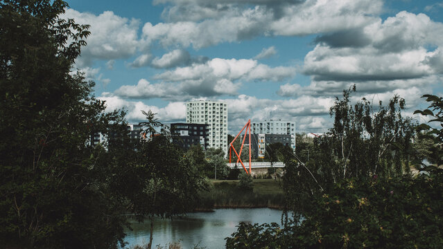 Contemporary urban landscape with residential buildings, bridge and calm water, framed by green trees under a cloudy sky.