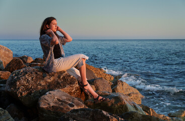 A girl poses and relaxes by the sea, sitting on rocks near the slope of a high cliff, panoramic view of the seashore and mountains at sunset