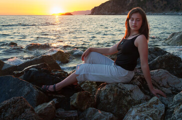 A girl poses and relaxes by the sea, on the rocks, a panoramic view of the coast and mountains at sunset, sunlight creates backlighting and silhouettes