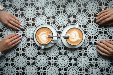 Top-down view of two cappuccino cups with latte art on a white lace tablecloth, featuring the hands...