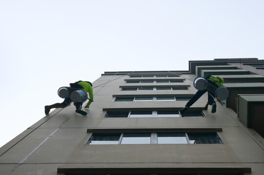 Two unrecognizable workers performing maintenance work high up on the exterior of a building, likely cleaning or applying sealant.