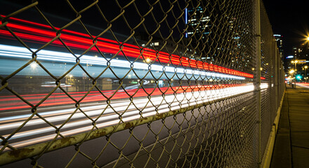 Long exposure shot through a chain-link fence showing red and white light trails from cars on a city highway at night