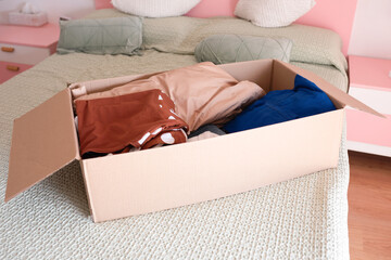 Cardboard box filled with assorted clothing items, placed on a neatly made bed, showcasing a cozy bedroom environment with soft textures and pastel colors, ideal for organization themes