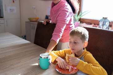 Young boy eating breakfast at the kitchen table while an adult places a drink nearby, morning daily routine and care.