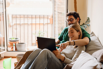 Man puts his arm around woman and points at laptop she holds on her lap while they smile and sit on bed
