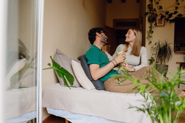 Obraz premium Woman puts her hand on man's shoulder while they sit on bed and hold cups while they laugh