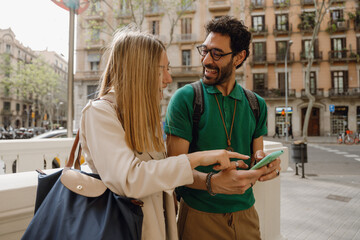 A woman points at a phone held by a man who is talking while they stand next to each other and laugh