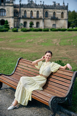 Elegant woman in a pale dress relaxing on a wooden bench in a park with a historic castle in the background, creating a calm portrait, soft light enhances fabric, poised posture, tranquil atmosphere,