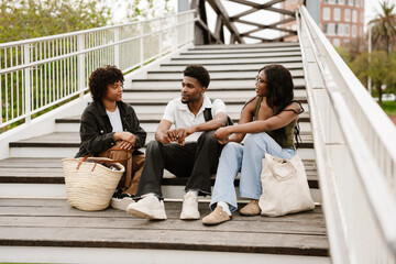A group of three friends sit on the steps of a bridge while one of them talks and one of them smiles