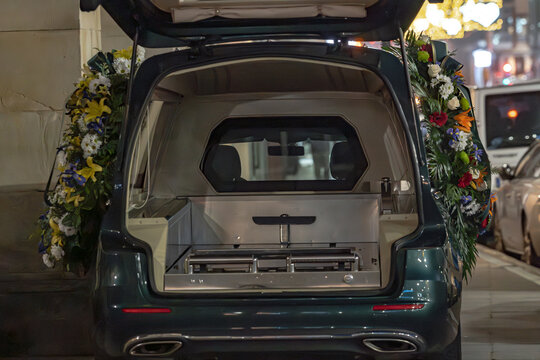 A close-up, rear view of a dark hearse with its tailgate open, revealing the polished interior and gurney. Large, colorful floral wreaths are attached to the sides, marking a somber occasion.