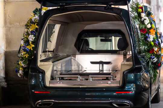 A close-up, rear view of a dark hearse with its tailgate open, revealing the polished interior and gurney. Large, colorful floral wreaths are attached to the sides, marking a somber occasion.