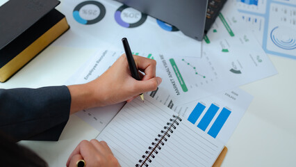 Businesswoman analyzing financial reports, holding a notebook and pen on modern office desk, focused on data charts, planning strategy, performance review, and corporate decision making.