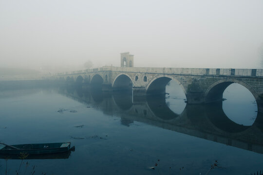Mecidiye bridge arches reflecting in meric river with a small boat floating in the serene, foggy morning, creating a historical and mystic atmosphere
