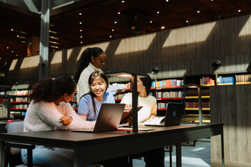 A group of three female students are sitting at a table in front of laptops and talking while a female student is standing next to them holding a notebook and they are laughing