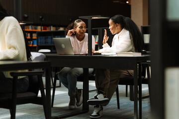 A female student gestures and points to a female student sitting next to her at a table in front of a laptop while they talk