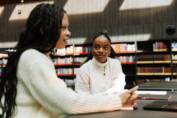 Female student listening to female student talking and holding pen over notebook while they sit at table