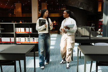 Female student holding notebooks and listening to female student holding laptop and talking while gesturing as they walk between desks