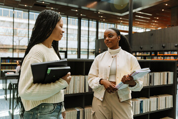 Two female students stand near bookshelves while one holds notebooks and the other a laptop © Drobot Dean