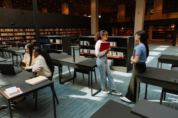 Two female students stand and talk while leaning on tables and two female students sit and talk next to them