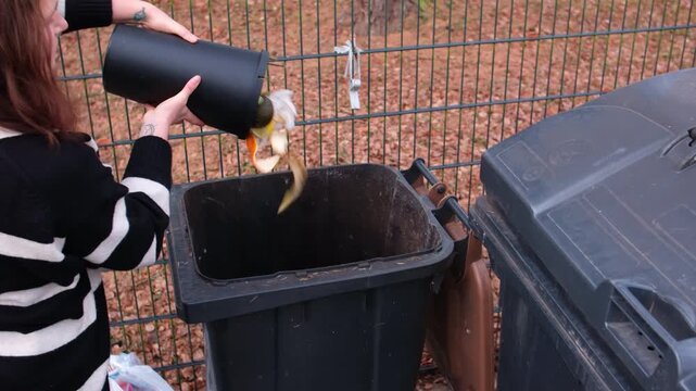 Young woman in striped sweater sorts organic food waste into the brown bio bin outdoors in Germany. Focus on correct trash separation, sustainability, and European lifestyle.