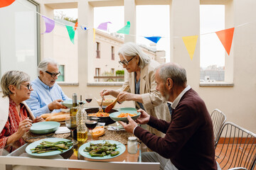 A woman stands and holds a fork and spatula over a frying pan while a group of three friends sit at a table next to her and hold plates