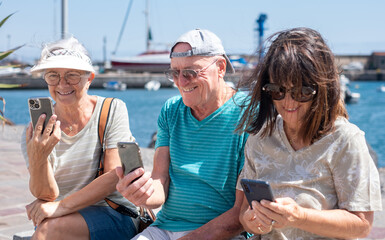 Group of three happy seniors in vacation looking at their smartphones enjoying a sunny day sitting near the harbor. A healthy and peaceful lifestyle in retirement, outdoors, by the sea.