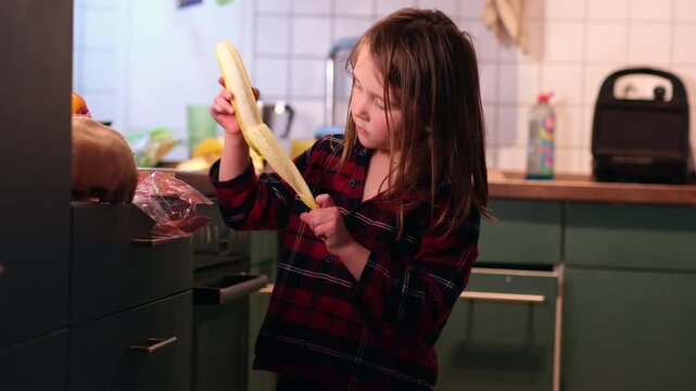 Cute Caucasian girl (age range, e.g., 4-6) in pajamas smiling while peeling a ripe banana in the bright kitchen. Healthy breakfast preparation, early morning, happy childhood moments at home.