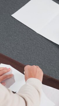hands pinning pattern to brown fabric edge, precision cutter adjusts paper guide and smooths seam allowance, tailor finalizes placement, finisher trims excess near green mockup, focused workspace