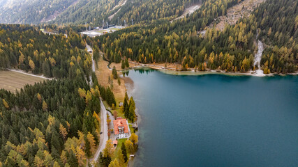 aerial landscape view of area along east coast of turquoise Antholzer See, a lake in Antholzertal in South Tyrol, Italy, with outflow and "Antholzer Wildsee Haus" hotel, surrounded by autumn forest