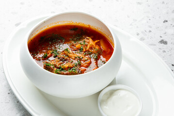 Appetizing photo of hot, rich Ukrainian/Russian borscht soup or hodgepodge (solyanka) with meat, cabbage, and herbs, served with sour cream on a white plate in a restaurant setting