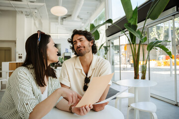 A man listens to a woman talking and points to a menu she is holding while they sit at a table