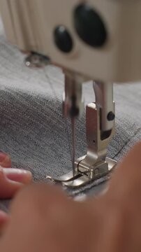 closeup asian seamstress sewing denim on industrial machine, fingers guiding gray fabric under presser foot, needle piercing cloth to create precise stitches, button detail visible, warm workshop