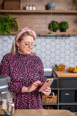 Woman in floral dress with pink headband is using smartphone in a modern kitchen, surrounded by fresh ingredients and kitchen appliances, showcasing culinary creativity and lifestyle