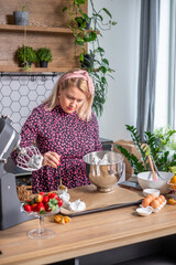 Woman in floral dress preparing dessert in modern kitchen, mixing ingredients in bowl, surrounded by fresh fruits and baking tools, showcasing culinary creativity and home cooking