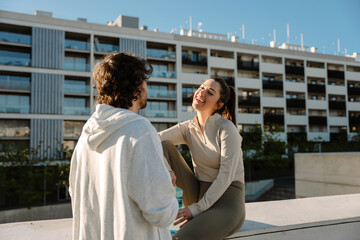 Fototapeta premium Female athlete sitting on a wall and laughing while holding a bottle and listening to a male athlete standing next to her
