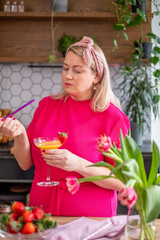 Fototapeta premium Woman in bright pink dress preparing a colorful cocktail in a modern kitchen, surrounded by fresh flowers and fruits, showcasing vibrant culinary creativity and style