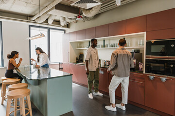 Fototapeta premium A male employee is talking to another male employee while they are standing at a table with two female employees standing and sitting behind it while one of them is looking at her phone