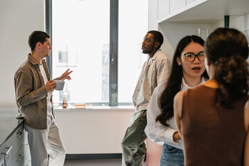 Fototapeta premium A male worker holds a cup and gestures while talking to a man across from him while two female workers stand next to them, one of whom is talking